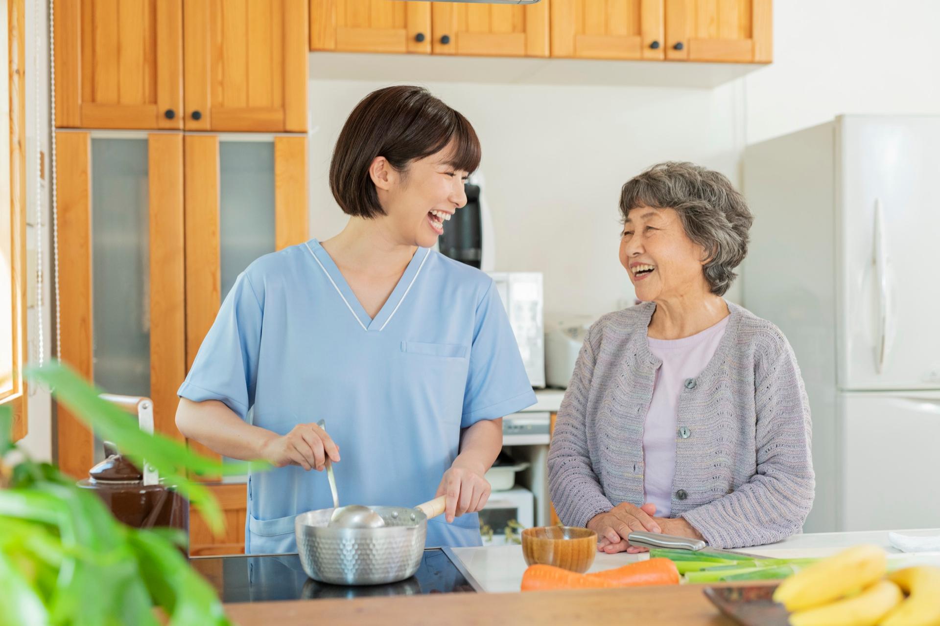 A caregiver and a senior woman laughing together while cooking a meal in a bright kitchen