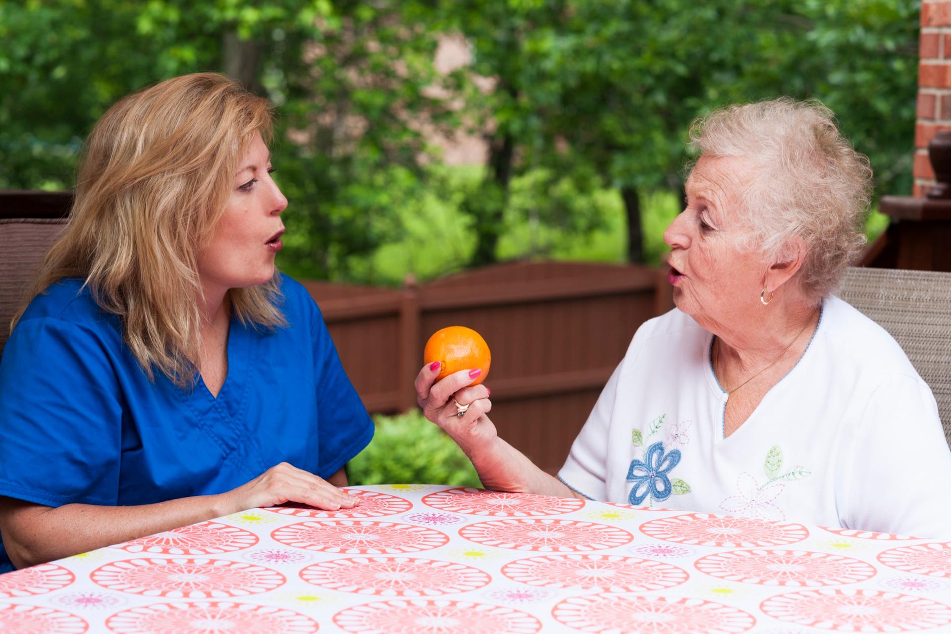A speech-language pathologist practicing word sounds with a senior woman at a patio table